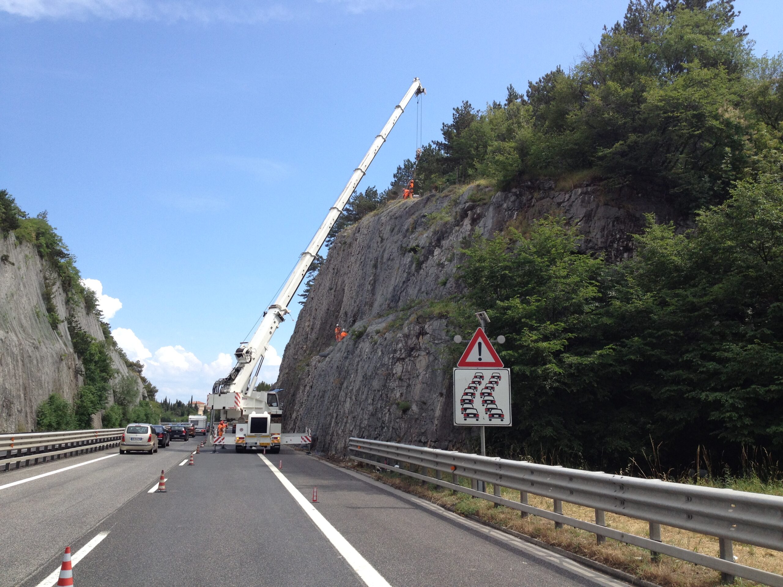 Coda in autostrada per lavori con autogrù e operai appesi per stendere reti paramassi su parete franosa